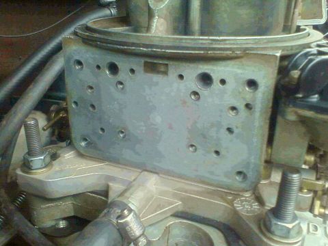 1981 Ford Bronco - Grainy close-up of the side of a carburetor's main body sitting on an aluminum intake manifold, showing the machined face drilled with nume…