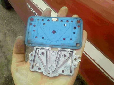1981 Ford Bronco - A greasy hand holds two carburetor metering blocks for comparison: a blue-painted cast block above and a bare aluminum block with machined…