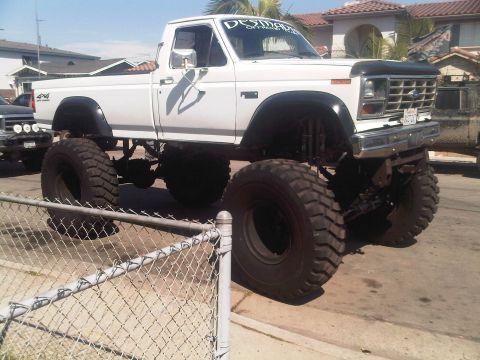 1986 Ford Bronco - A heavily modified white pickup truck with oversized tires is parked on a street, with another vehicle visible in the background.