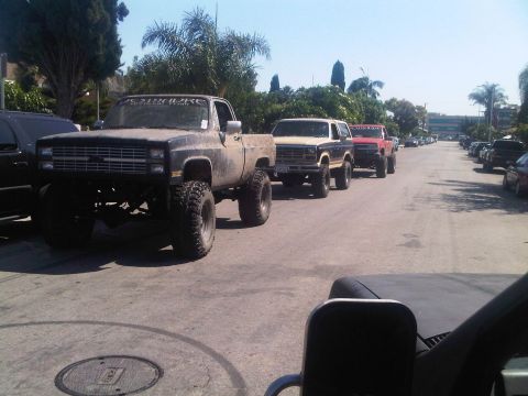 1986 Ford Bronco - Several modified trucks and SUVs are parked in a line on a street.