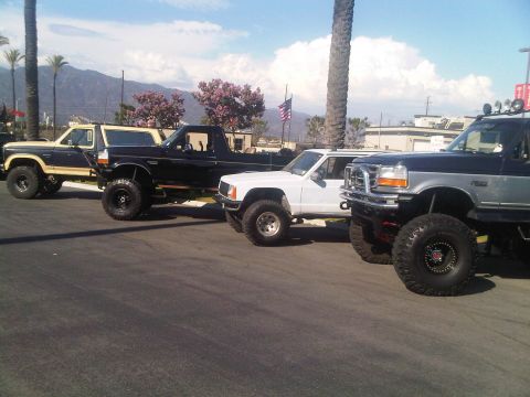 1986 Ford Bronco - Several modified trucks are parked in a lot under a bright sky with mountains in the background.