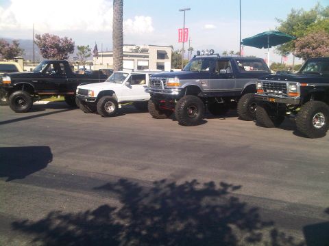 1986 Ford Bronco - Several modified trucks and SUVs are lined up in a parking lot under a bright sky.