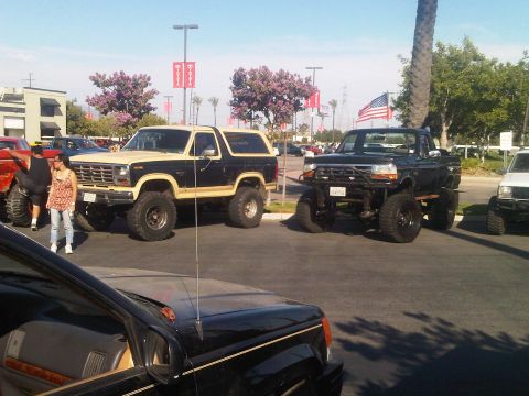 1986 Ford Bronco - A lifted two-tone Ford Bronco and a lifted black Ford pickup truck are parked at an outdoor event.