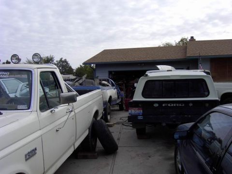 1980 Ford Bronco - Several vehicles are parked in a driveway in front of a garage, with one truck elevated on blocks.