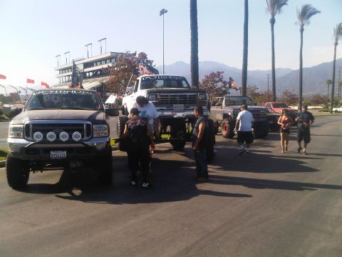 1986 Ford Bronco - A collection of modified trucks and SUVs are gathered in a parking lot, with a race track grandstand visible in the distance.