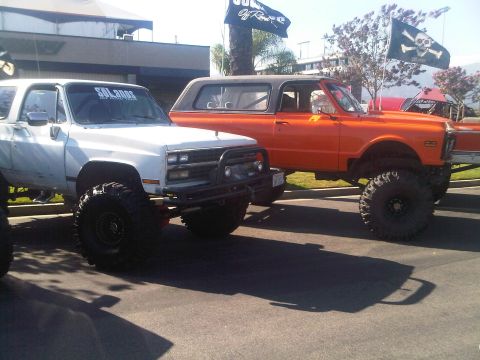 1986 Ford Bronco - Two lifted SUVs, one white and one orange, are parked outdoors with flags visible.