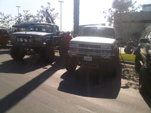 1986 Ford Bronco - Two heavily modified trucks are parked side-by-side in a lot, with a third vehicle partially visible on the right.
