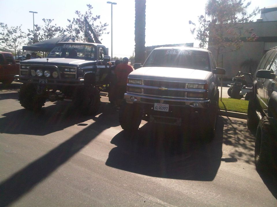 Two lifted pickup trucks parked outside, one with a flag on top