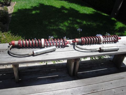 1986 Ford Bronco - Two red coilover shock absorbers with silver reservoirs are displayed outdoors on a wooden table.