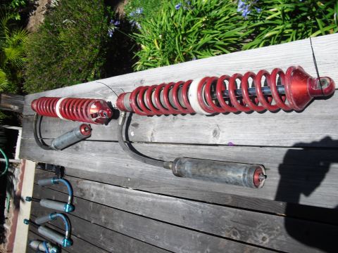 1986 Ford Bronco - Two red coilover shock absorbers with external reservoirs are laid out on a wooden surface, likely for an off-road vehicle.