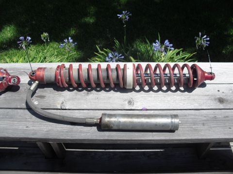 1986 Ford Bronco - A red coilover shock absorber with an external reservoir is displayed on a wooden surface, with a grassy, flowered background.