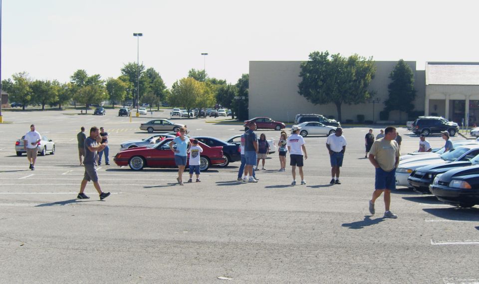 People milling around a parking lot with several cars, including a red sports car