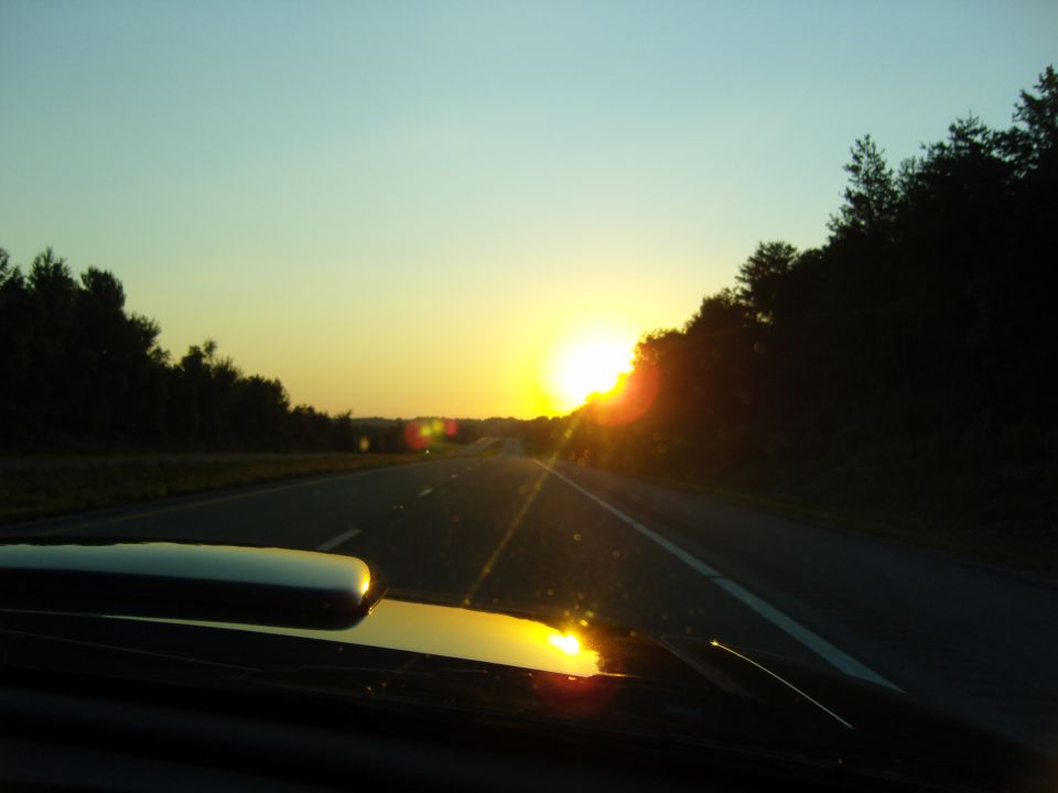 View from inside a car looking out at a highway during sunset, with trees lining the road