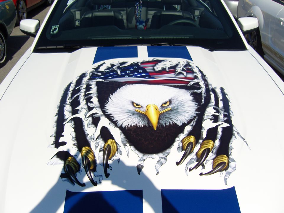 White Ford Mustang hood with blue racing stripes and a graphic of an eagle tearing through the paint, showing the American flag underneath. Another white car is visible to the right