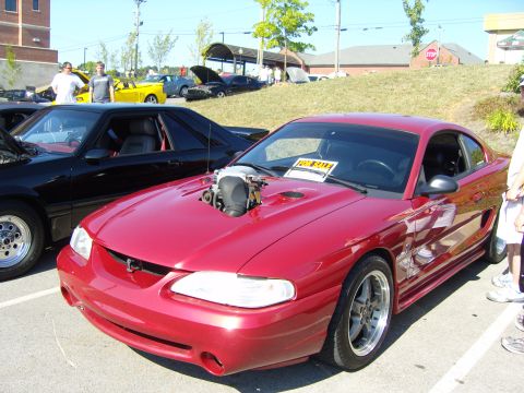 2007 Ford Mustang - A red Ford Mustang with a modified engine protruding from the hood is parked at an outdoor car show.