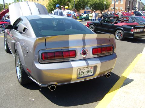 2007 Ford Mustang - A silver Ford Mustang with racing stripes is parked at a car show, with other vehicles visible in the background.