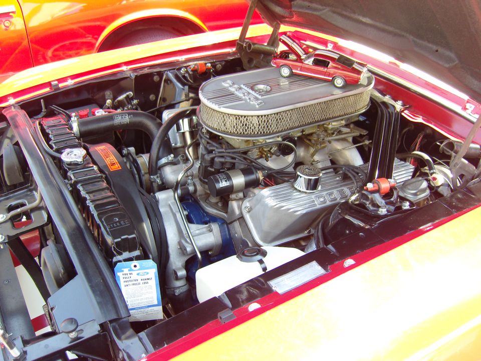 Red 1969 Ford Mustang engine bay with a toy car on the air cleaner. Another red car is visible in the background