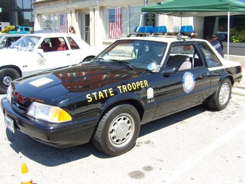 2007 Ford Mustang - A 1987 Ford Mustang police car is parked next to another vehicle, with American flags visible in the background.