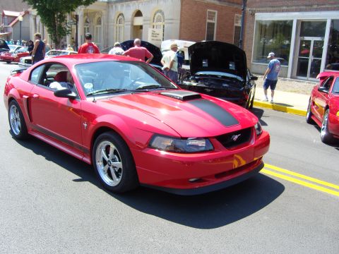2007 Ford Mustang - A red 2007 Ford Mustang is parked on a street, with other vehicles and people in the background.