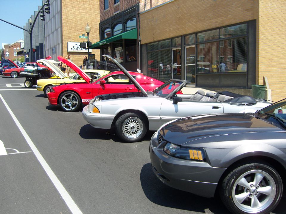 Several Ford Mustangs parked on a street, some with hoods up. The closest one is a dark gray model