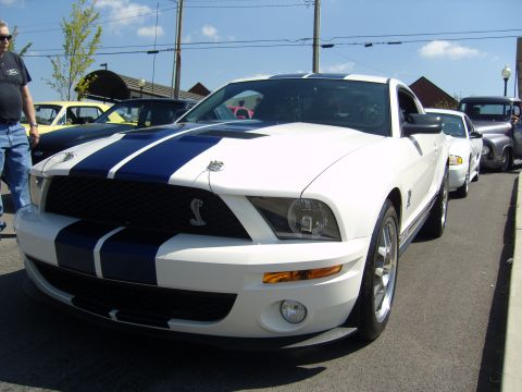 2007 Ford Mustang - A white 2007 Ford Mustang Shelby GT500 with iconic blue racing stripes is parked in a line with other vehicles.