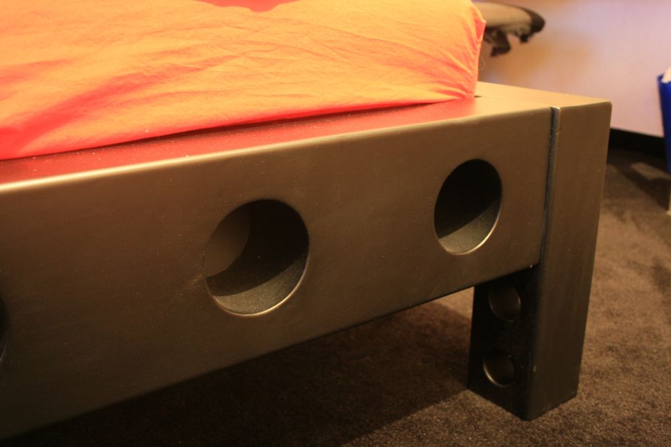 Close-up of a dark metal bed frame with circular cutouts and an orange blanket