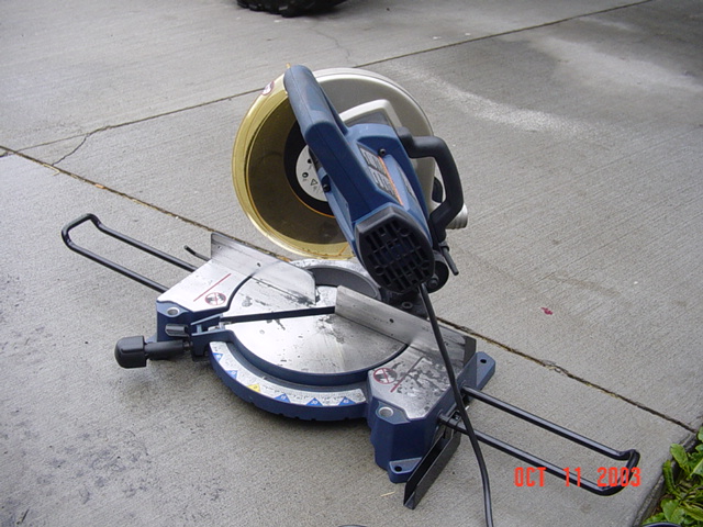 Blue compound miter saw sitting on a concrete driveway, power cord coiled