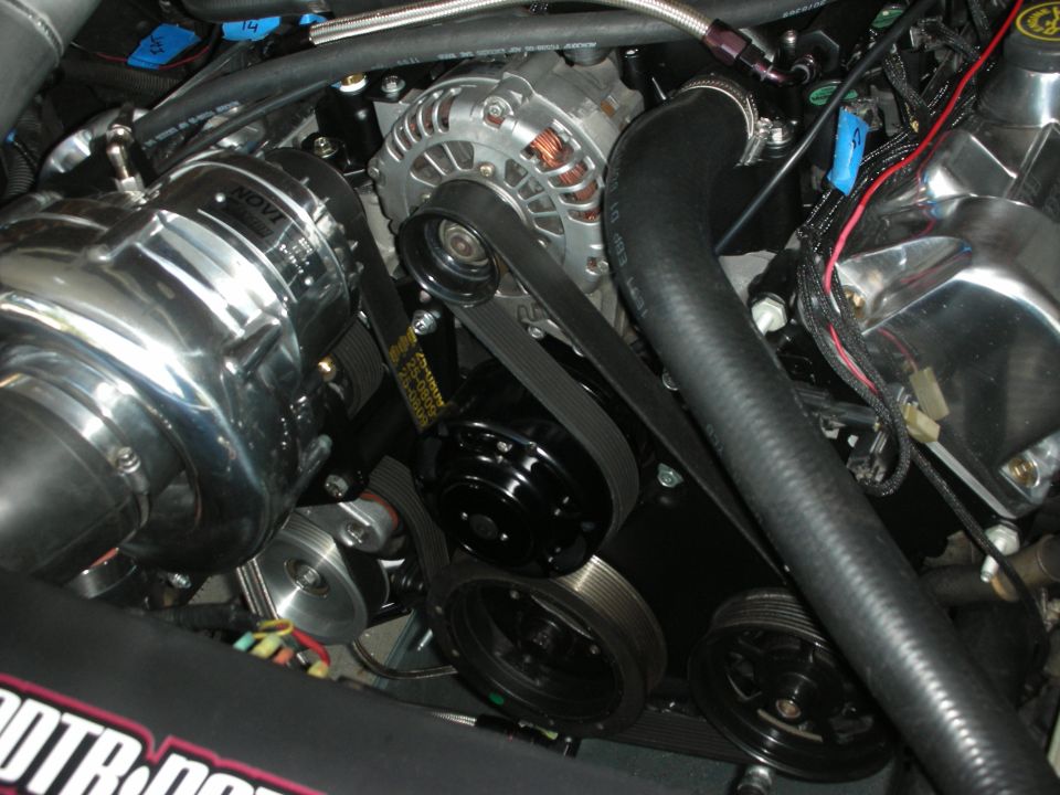 Engine bay with a polished Novi supercharger, alternator, and serpentine belt system. Some wiring and hoses are visible