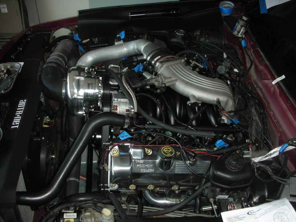 Engine bay with a polished supercharger, intake manifold, and valve covers. Some wiring is taped up
