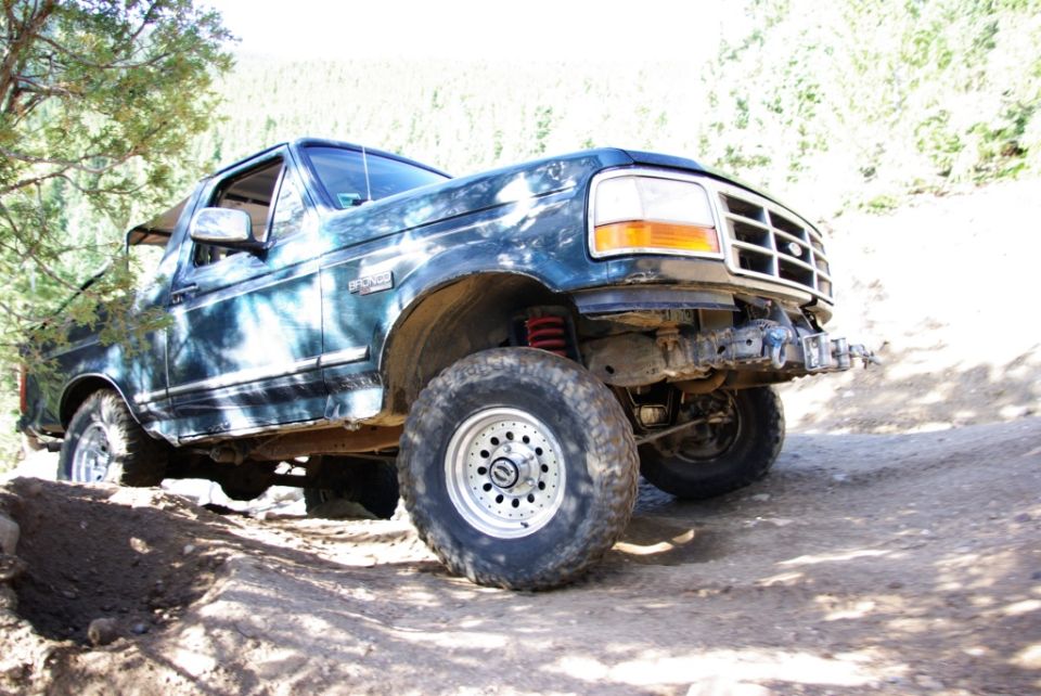 Dark blue Ford Bronco on a dirt trail, front bumper removed, lifted suspension