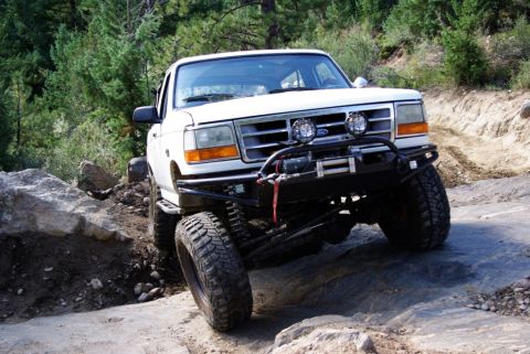 1995 Ford Bronco - White Ford Bronco tackles a challenging off-road course with large tires and modified suspension.