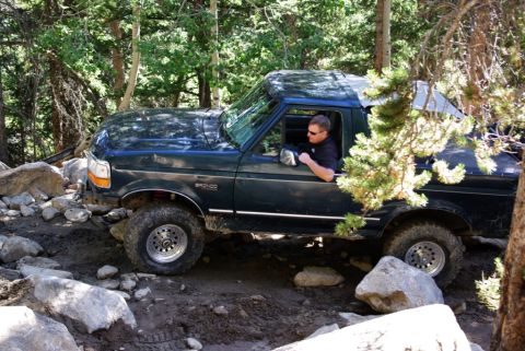 1995 Ford Bronco - A dark green Ford Bronco is driven off-road through a rocky, wooded area.