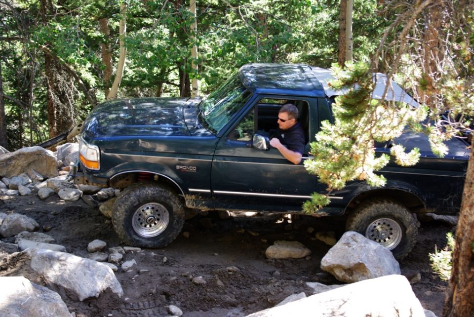 Dark green Ford Bronco navigating a rocky, wooded trail. Driver wears sunglasses
