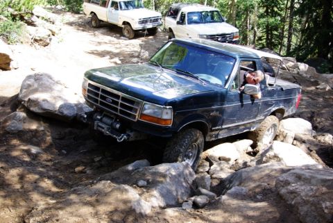 1995 Ford Bronco - A dark blue Ford F-Series truck is shown navigating a difficult, rocky off-road trail with two other vehicles following.