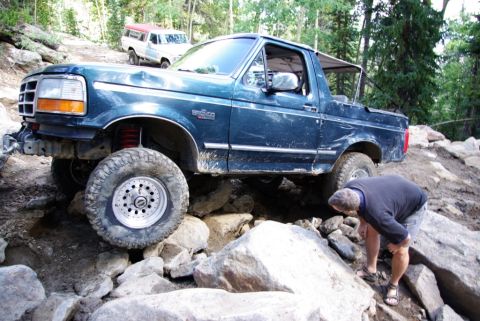 1995 Ford Bronco - A dark blue Ford Bronco is shown off-roading on a rocky trail, with another vehicle visible in the background.