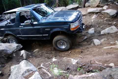 1995 Ford Bronco - 1995 Ford Bronco tackling a challenging rocky trail with trees and boulders visible.
