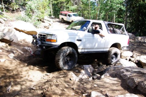 1995 Ford Bronco - A white 1995 Ford Bronco tackles a challenging off-road path, with a second vehicle visible in the background.