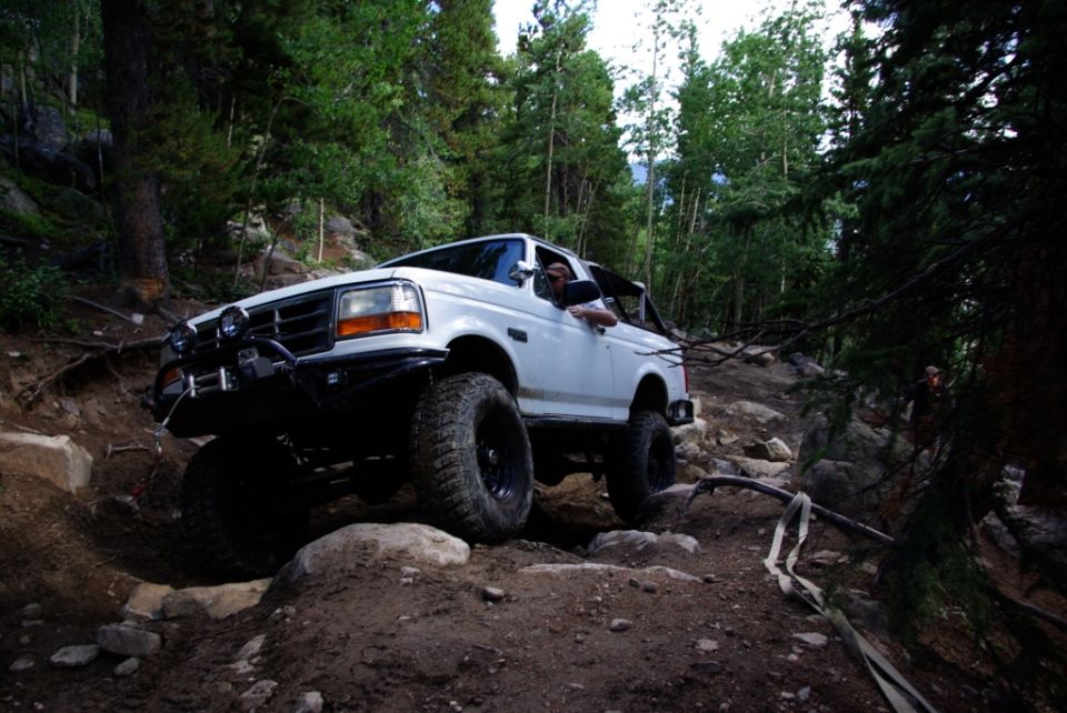 White 1995 Ford Bronco driving on a rocky, wooded trail. Driver's side window is down
