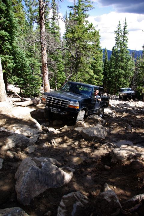 1995 Ford Bronco - A 1995 Ford Bronco navigates a challenging rocky trail in a wooded area, with another vehicle visible further down the path.