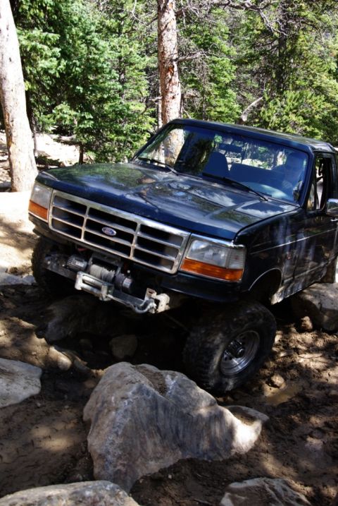 1995 Ford Bronco - A Ford Bronco navigates a challenging, rocky off-road path surrounded by trees.