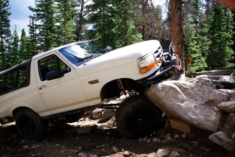 1995 Ford Bronco - A white 1995 Ford Bronco is shown off-roading over large rocks on a wooded trail.
