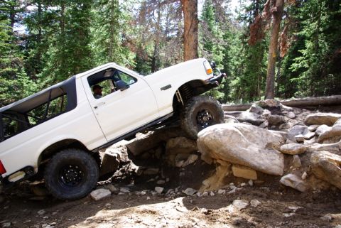 1995 Ford Bronco - A white 1995 Ford Bronco tackles a challenging rocky trail in a wooded area.