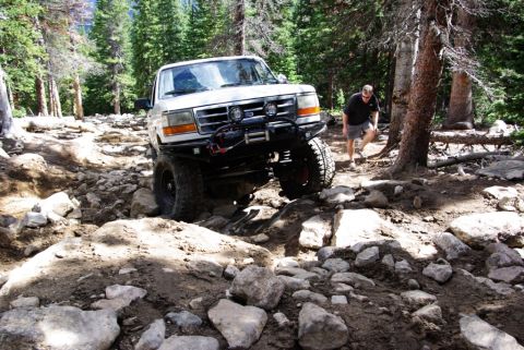 1995 Ford Bronco - A 1995 Ford Bronco tackles a challenging off-road path through a wooded area with large rocks.