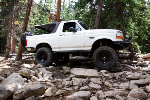 1995 Ford Bronco - A modified white Ford Bronco tackles a challenging rocky terrain in a wooded area.