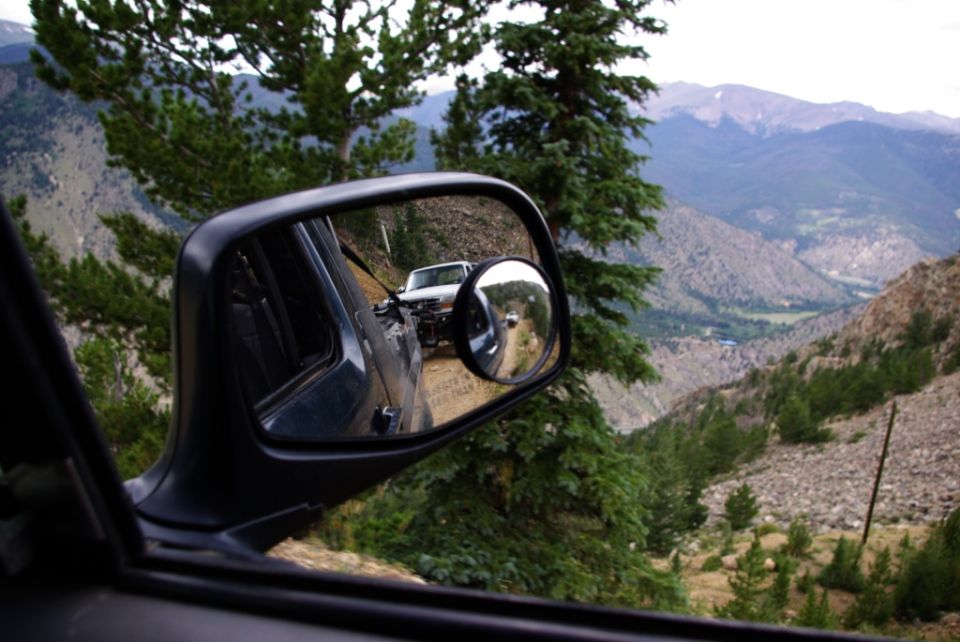 View in side mirror shows a white SUV on a dirt road in a mountainous landscape