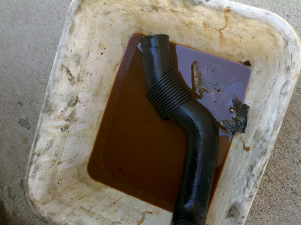Black corrugated hose draining brown fluid into white drain pan on concrete