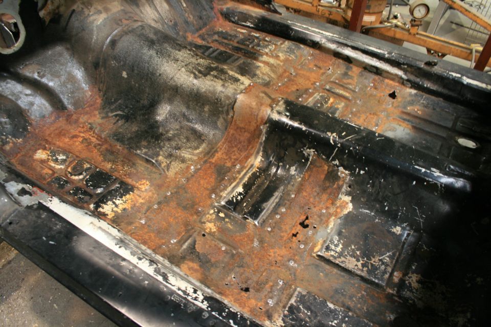 Black vehicle floor pan with extensive rust damage and missing sections, viewed from above