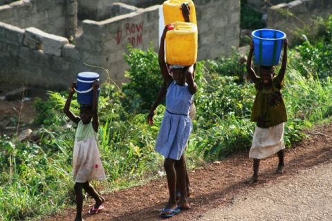 1987 Ford Bronco - Three children walk along a dirt road carrying water containers on their heads, with partially constructed buildings in the background.