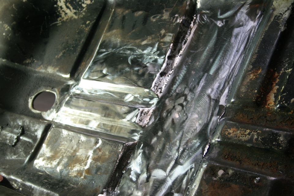 Undercarriage of a vehicle undergoing repair, showing metal floor pan and structural supports