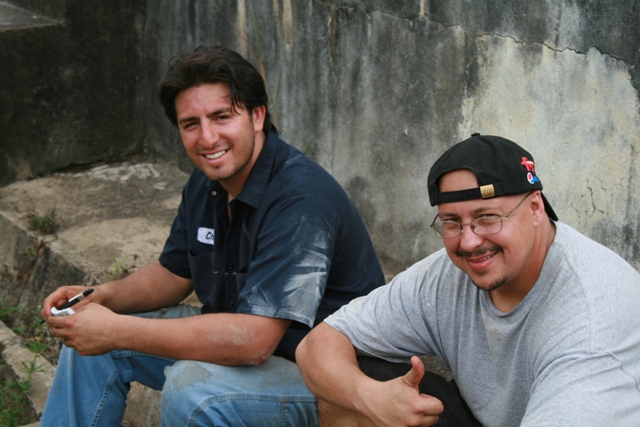 Two men sitting on concrete steps outdoors. One wears a blue work shirt, the other a grey t-shirt and baseball cap